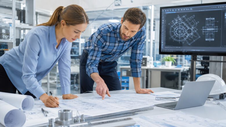 مهندسان يراجعان مخططات هندسية مطبوعة ورسمًا ميكانيكيًا معروضًا على برنامج أوتوكاد